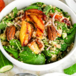 Quinoa salad bowl with grilled peach slices, candied pecans, spinach, kale, cucumber, and red onion.