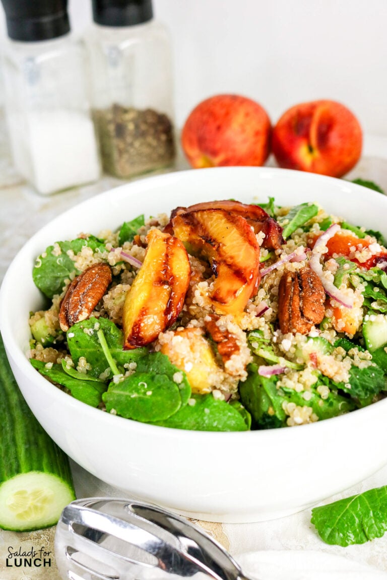 Quinoa salad bowl with grilled peach slices, spinach, pecans, cucumber, and red onion, with fresh peaches in the background.