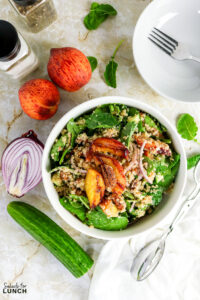 Quinoa salad bowl with grilled peach slices, arugula, cucumber, and red onion, surrounded by fresh peaches, cucumber, and red onion.