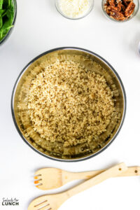 Cooked quinoa in a large stainless steel bowl, with spinach, shredded parmesan, and pecans in small bowls nearby.