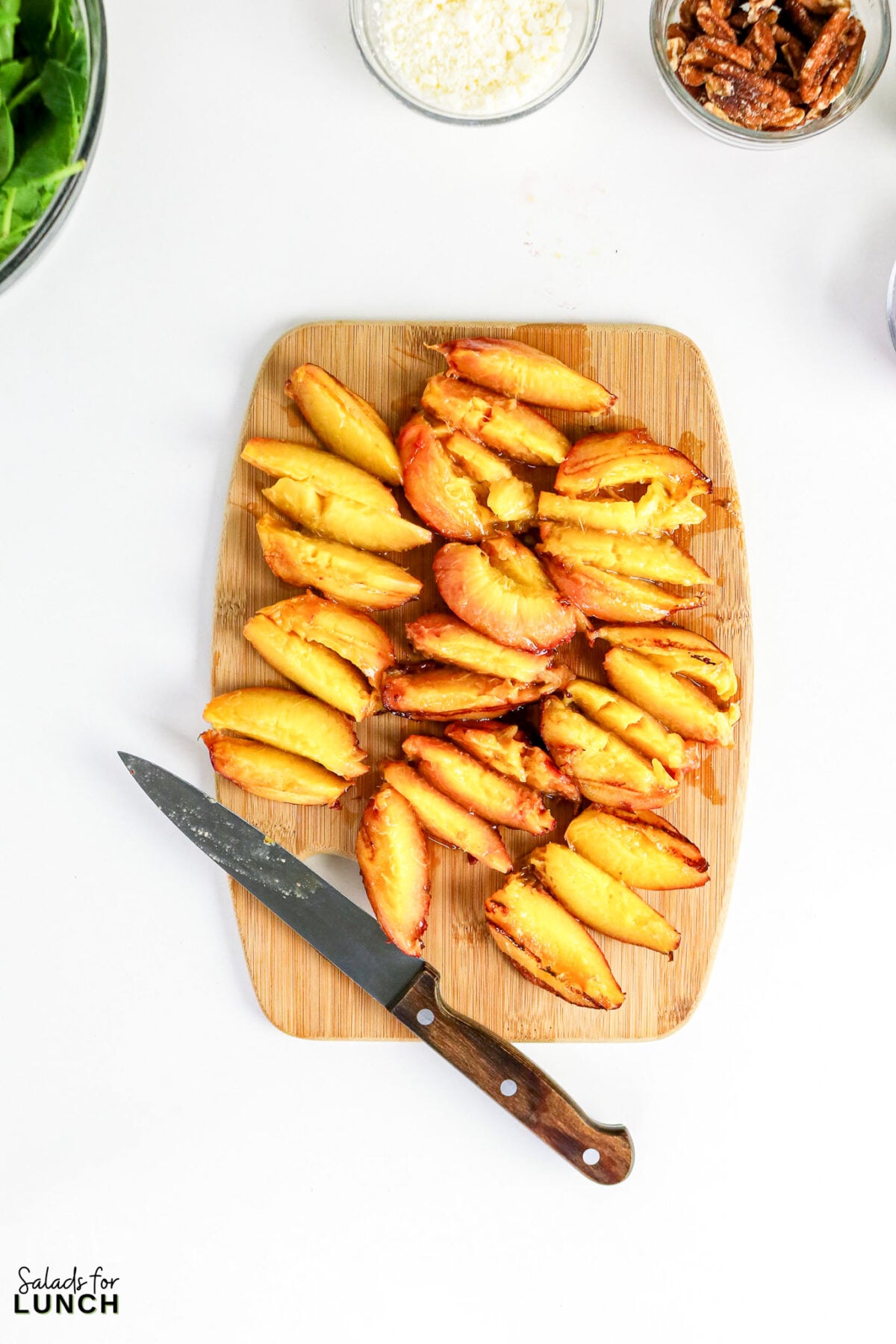 Sliced roasted peaches on a wooden cutting board with a knife, surrounded by bowls of spinach, shredded cheese, and pecans.