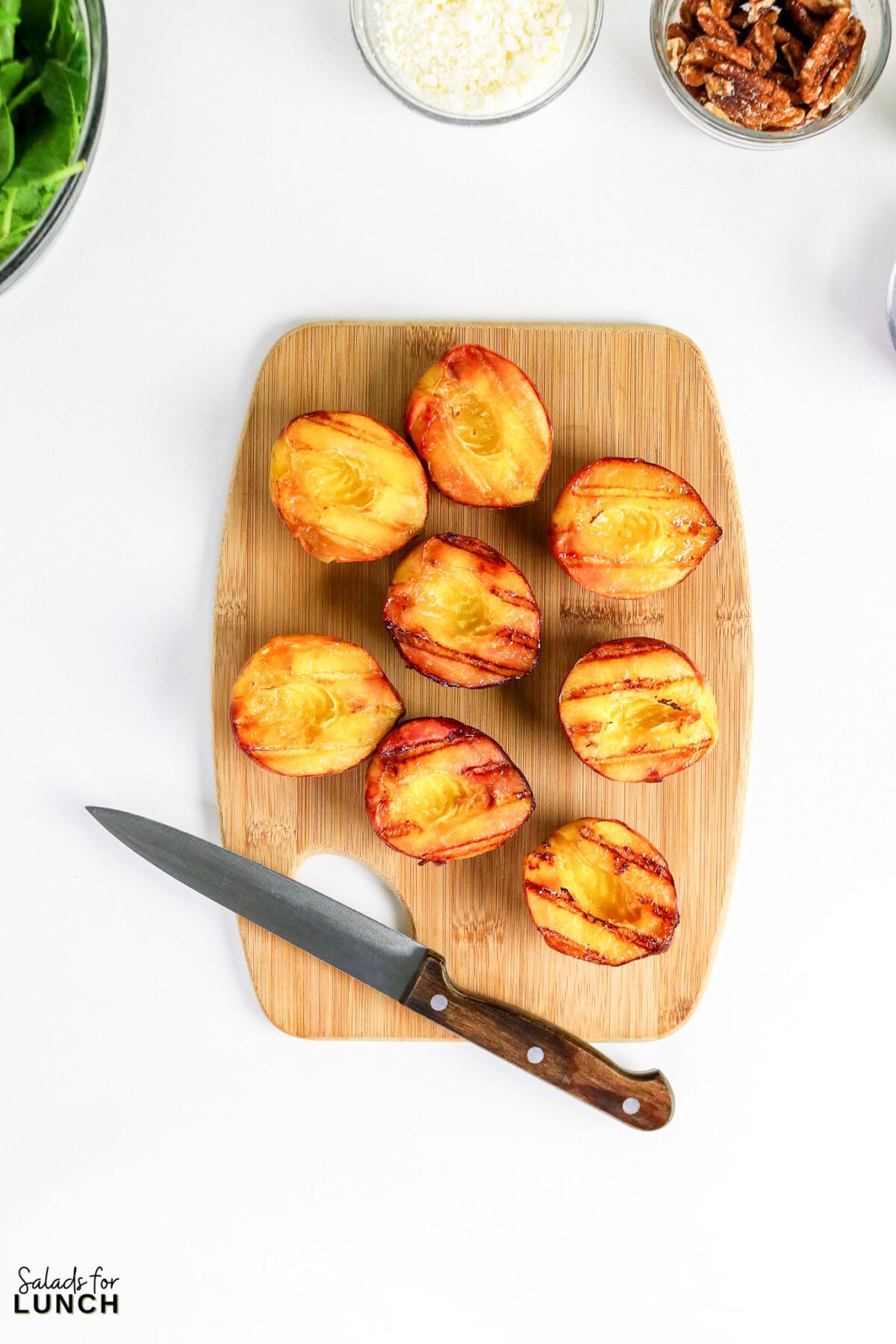 Grilled peach halves on a wooden cutting board with a knife, surrounded by bowls of spinach, cheese, and pecans.