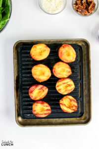 Eight grilled peach halves with char marks on a cast iron grill pan, with bowls of greens, cheese, and pecans nearby.