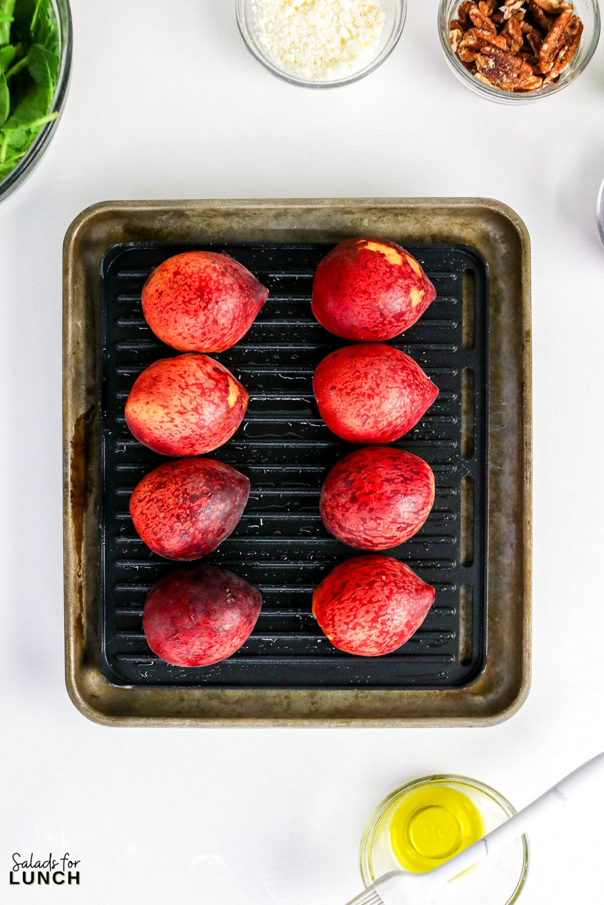 Eight peaches arranged on a grill pan, with bowls of arugual, cheese, pecans, and olive oil nearby.