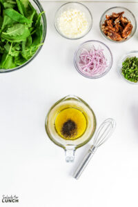 Salad ingredients laid out in glass bowls: arugula, feta cheese, pecans, sliced red onion, fresh herbs, and olive oil dressing with a whisk.