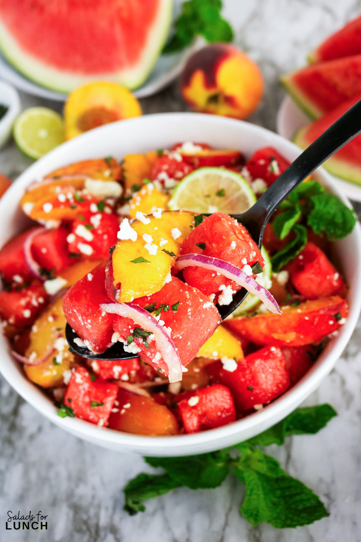 Bowl of watermelon peach salad with feta, red onion, mint, and lime, served with a black spoon.