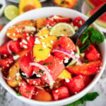 Bowl of watermelon peach salad with feta, red onion, mint, and lime, served with a black spoon.