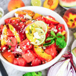 Watermelon peach salad with crumbled feta, red onion, mint, and a lime slice in a white bowl.