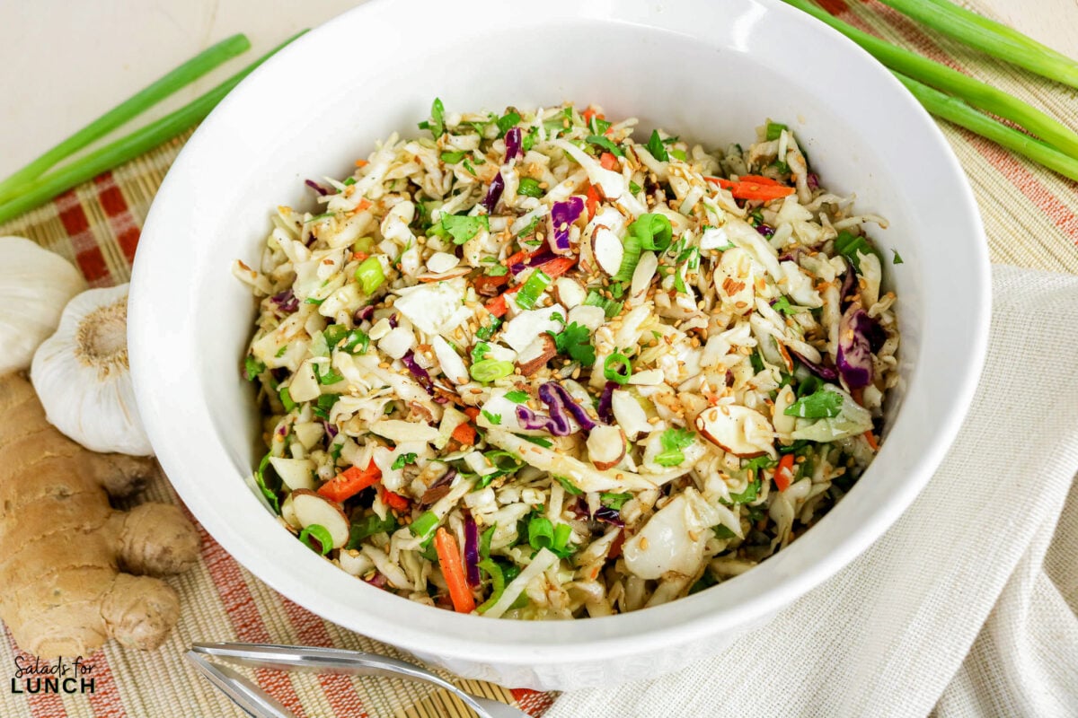 Asian coleslaw with shredded cabbage, carrots, sliced almonds, sesame seeds, and green onions in a white bowl.