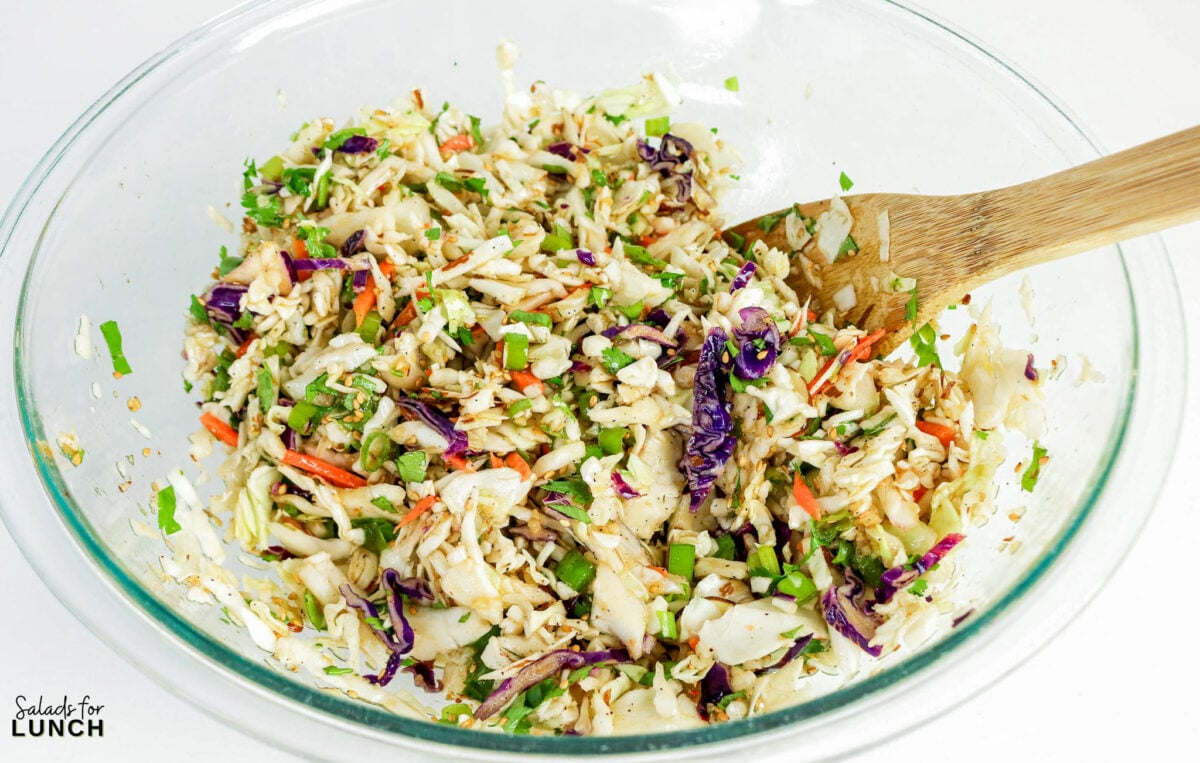 Asian coleslaw tossed with sesame ginger dressing in a glass bowl with a wooden spoon.