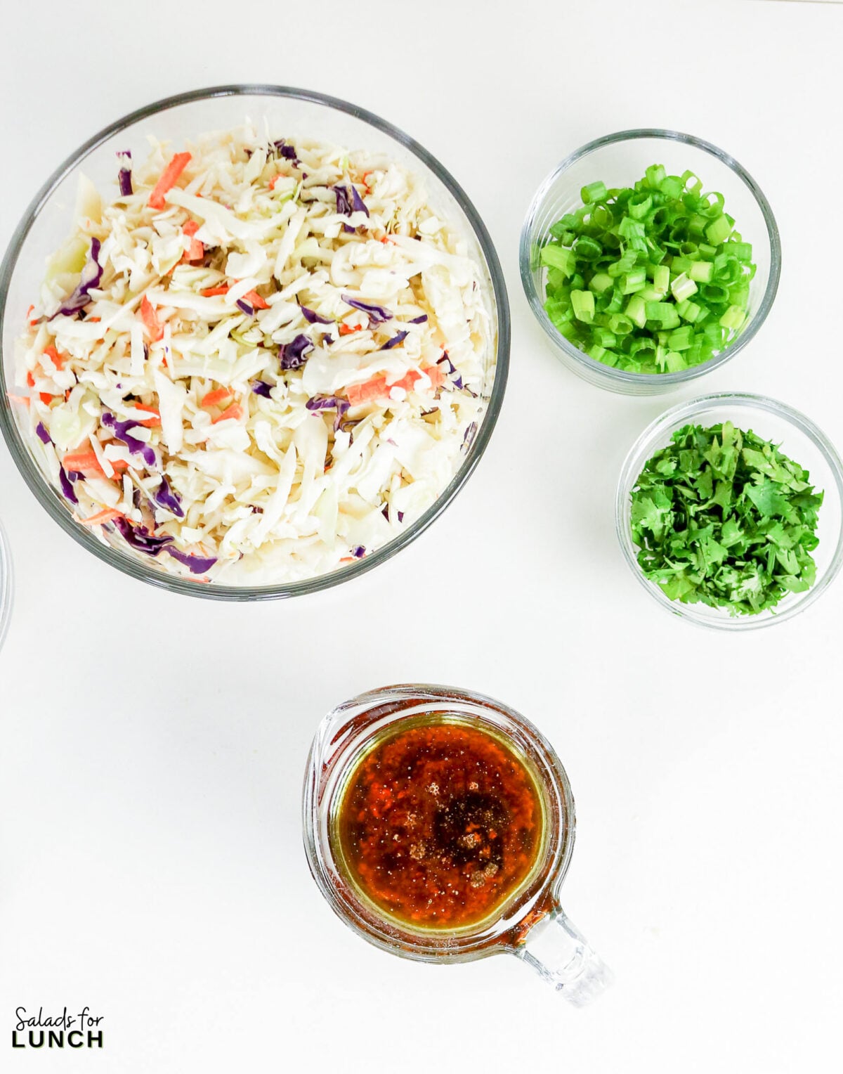 Overhead view of coleslaw mix, chopped green onions, cilantro, and Asian-style dressing in glass bowls.