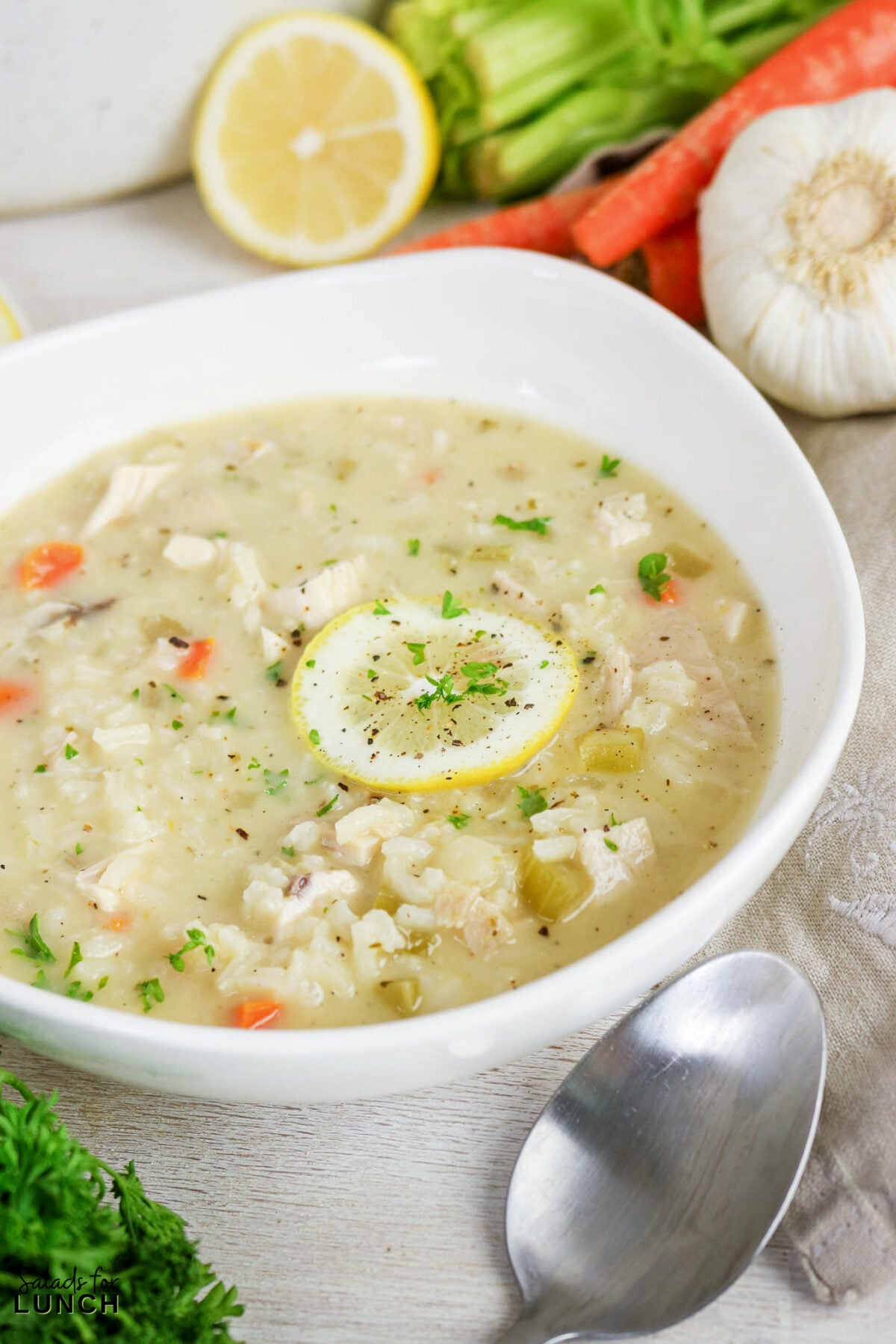 Greek Lemon Chicken and Rice Soup (Easy Avgolemono Recipe) - Greek Lemon Chicken Soup with Rice Recipe 12 Creamy Greek chicken and rice soup in a white bowl, garnished with a lemon slice and fresh parsley, with carrots and garlic in the background.