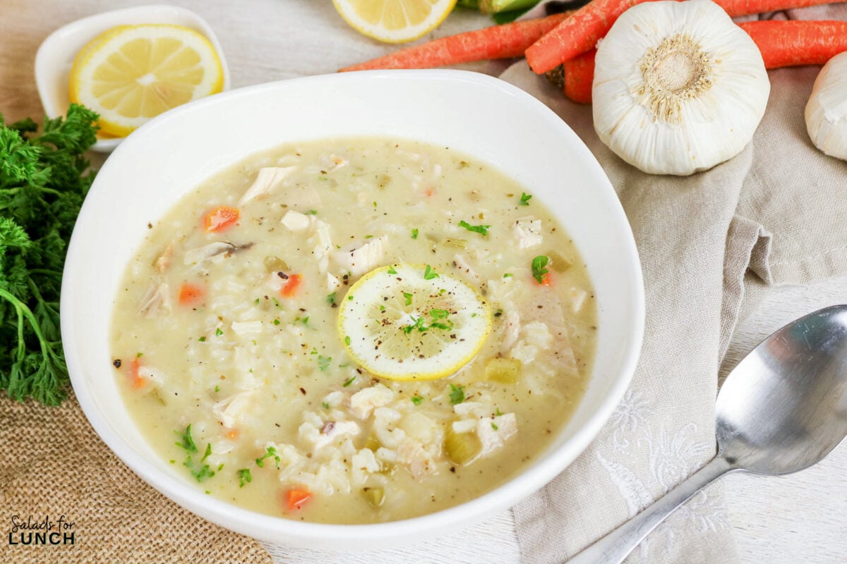 Greek Lemon Chicken and Rice Soup (Easy Avgolemono Recipe) - Greek Lemon Chicken Soup with Rice Recipe 13 Bowl of Greek chicken and rice soup garnished with a lemon slice and parsley, surrounded by fresh carrots, garlic, and lemon.