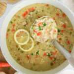 Overhead view of a pot of creamy chicken and rice soup with carrots, celery, and lemon slices, with a ladle scooping a serving.