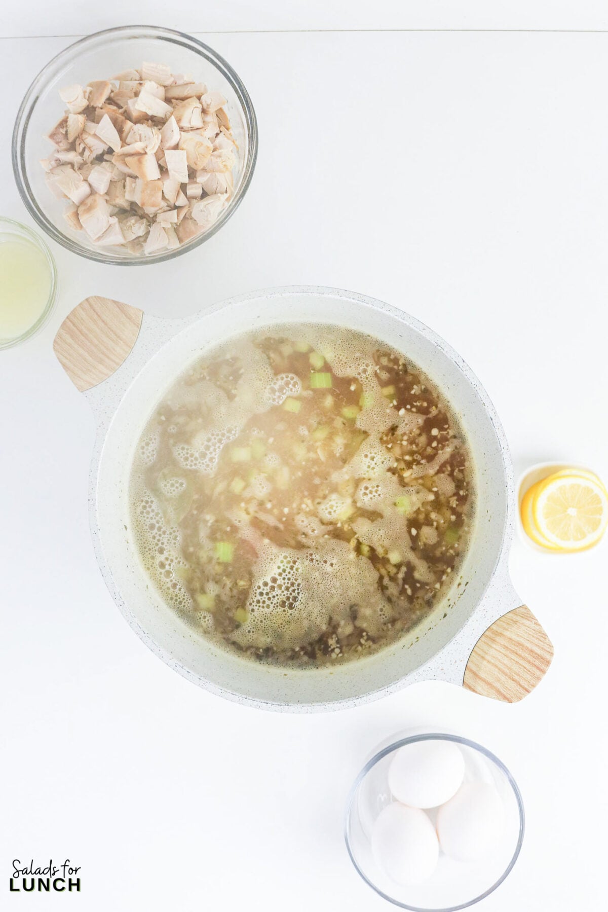 Greek Lemon Chicken and Rice Soup (Easy Avgolemono Recipe) - Greek Lemon Chicken Soup with Rice Recipe 35 Rice simmering in chicken broth with vegetables, surrounded by bowls of cooked chicken, eggs, lemon juice, and a halved lemon.