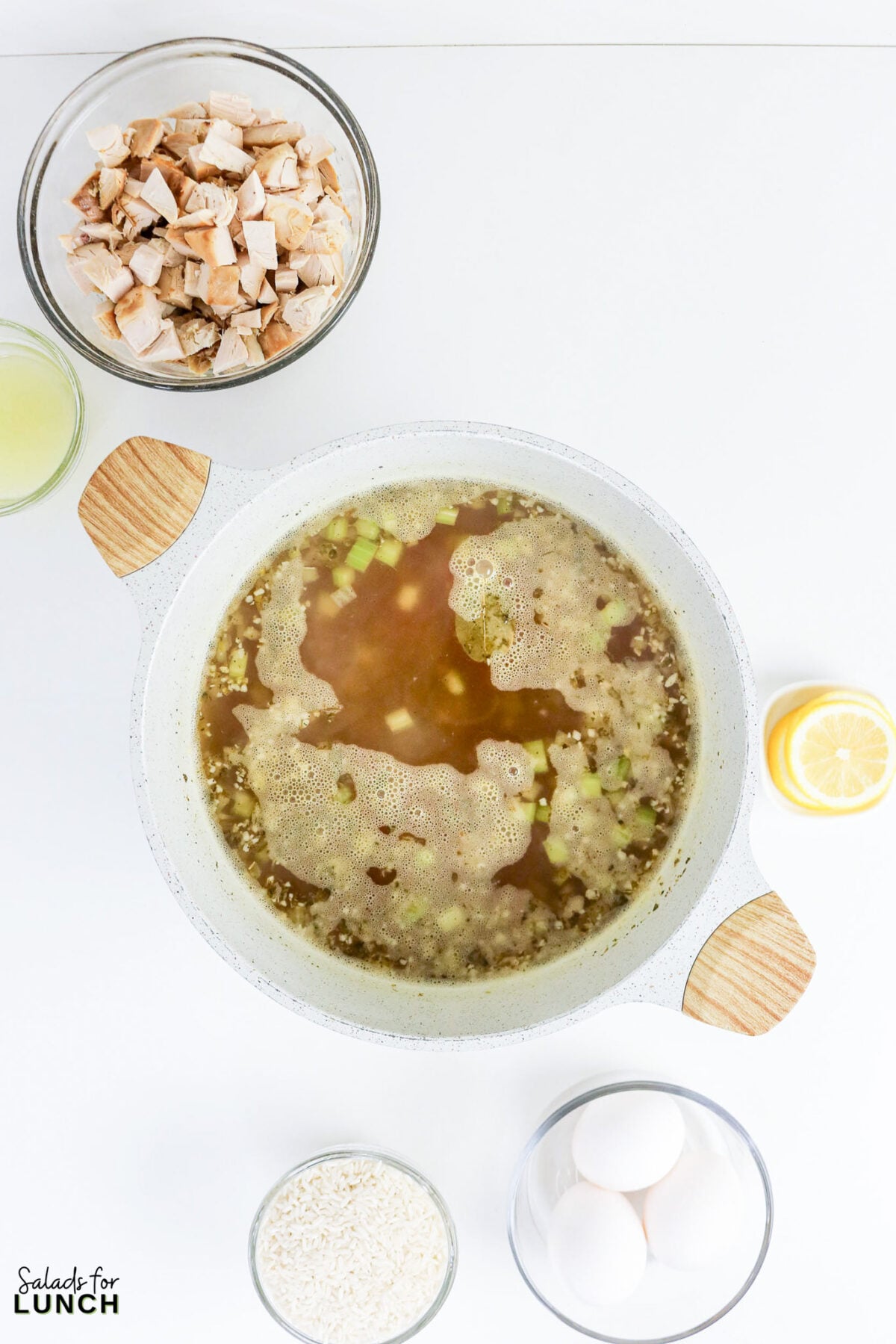 Greek Lemon Chicken and Rice Soup (Easy Avgolemono Recipe) - Greek Lemon Chicken Soup with Rice Recipe 36 Chicken broth with vegetables and a bay leaf boiling in a pot, surrounded by bowls of cooked chicken, rice, eggs, and lemon.