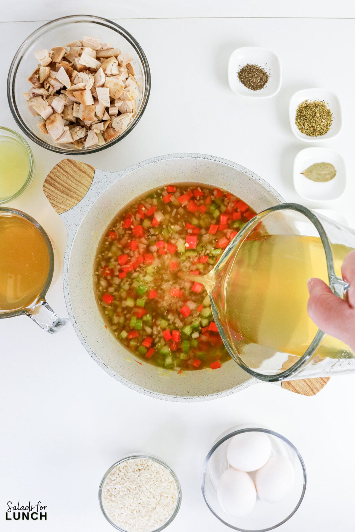 Greek Lemon Chicken and Rice Soup (Easy Avgolemono Recipe) - Greek Lemon Chicken Soup with Rice Recipe 39 Pouring chicken broth into a pot of sautéed vegetables, with cooked chicken, rice, eggs, and spices arranged nearby.