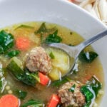 A bowl of Italian wedding soup with meatballs, dark leafy greens, and carrots, with a spoonful lifted above the bowl.