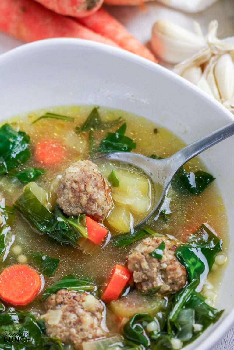 A bowl of Italian wedding soup with meatballs, dark leafy greens, and carrots, with a spoonful lifted above the bowl.