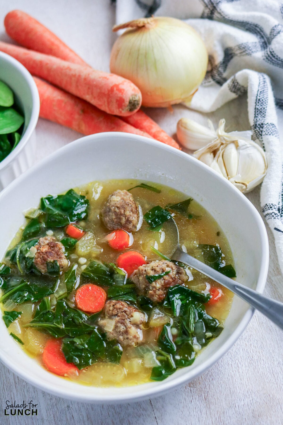 A bowl of Italian wedding soup with mini meatballs, wilted greens, carrots, and tiny pasta in light chicken broth, with fresh carrots, onion, and garlic alongside.