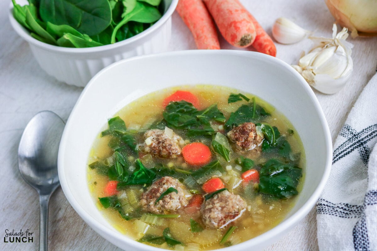 Bowl of Italian wedding soup with mini meatballs, wilted spinach, and carrot slices in chicken broth, with fresh ingredients in the background.
