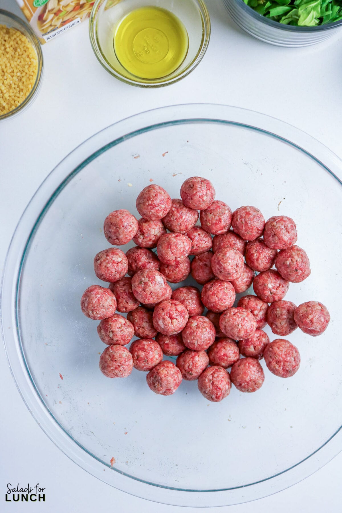 Raw 1-inch mini meatballs rolled and set aside in a glass bowl, with olive oil, tiny pasta, and greens in the background.