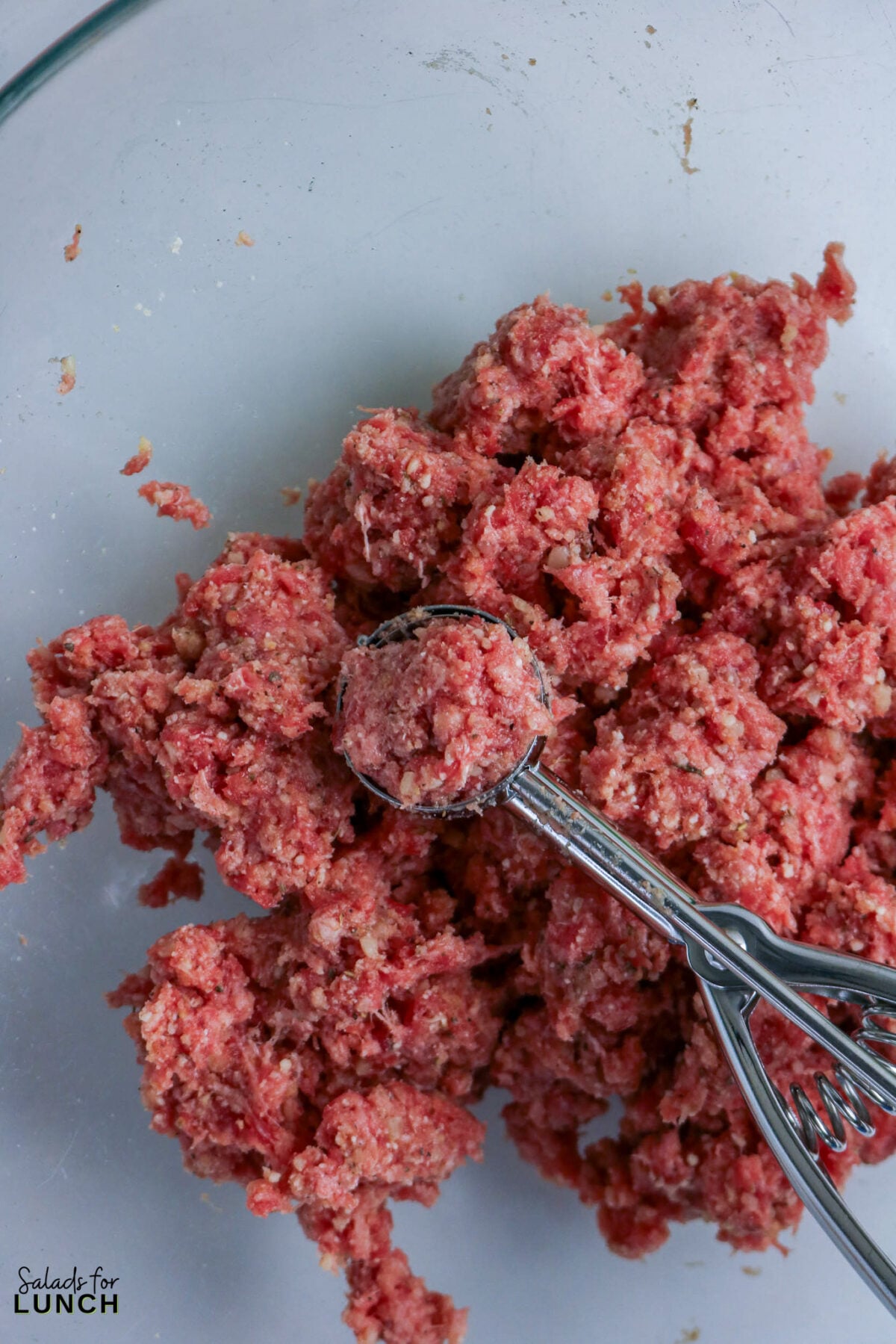 A cookie scoop portioning raw meatball mixture from a glass bowl to form 1-inch meatballs.