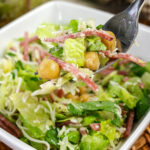A forkful of chopped Italian salad with romaine lettuce, salami, chickpeas, and shredded cheese held over a white serving bowl.