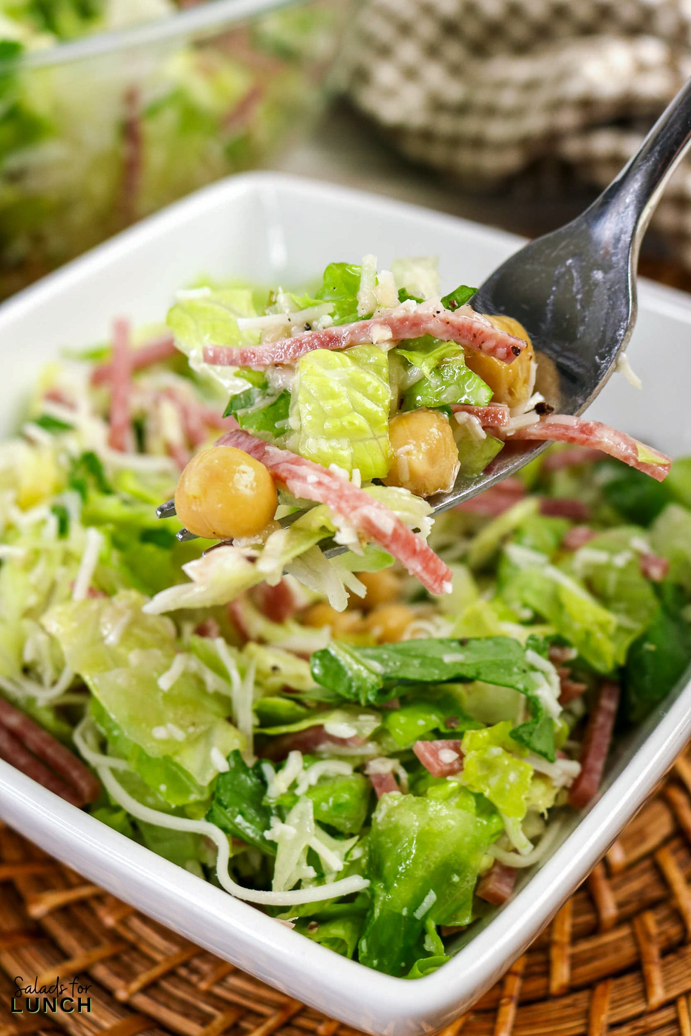 A forkful of chopped Italian salad with romaine lettuce, salami, chickpeas, and shredded cheese held over a white serving bowl.
