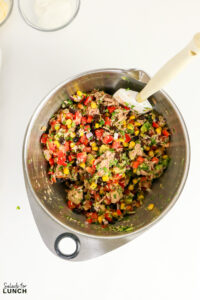 Tuna salad with corn, black beans, diced red pepper, avocado, and herbs mixed in a large stainless steel bowl with a spatula.