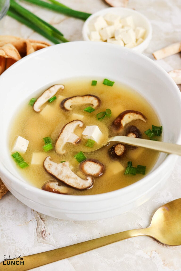 A white bowl of miso soup with shiitake mushrooms, tofu cubes, and sliced green onions, with a gold spoon resting in it.