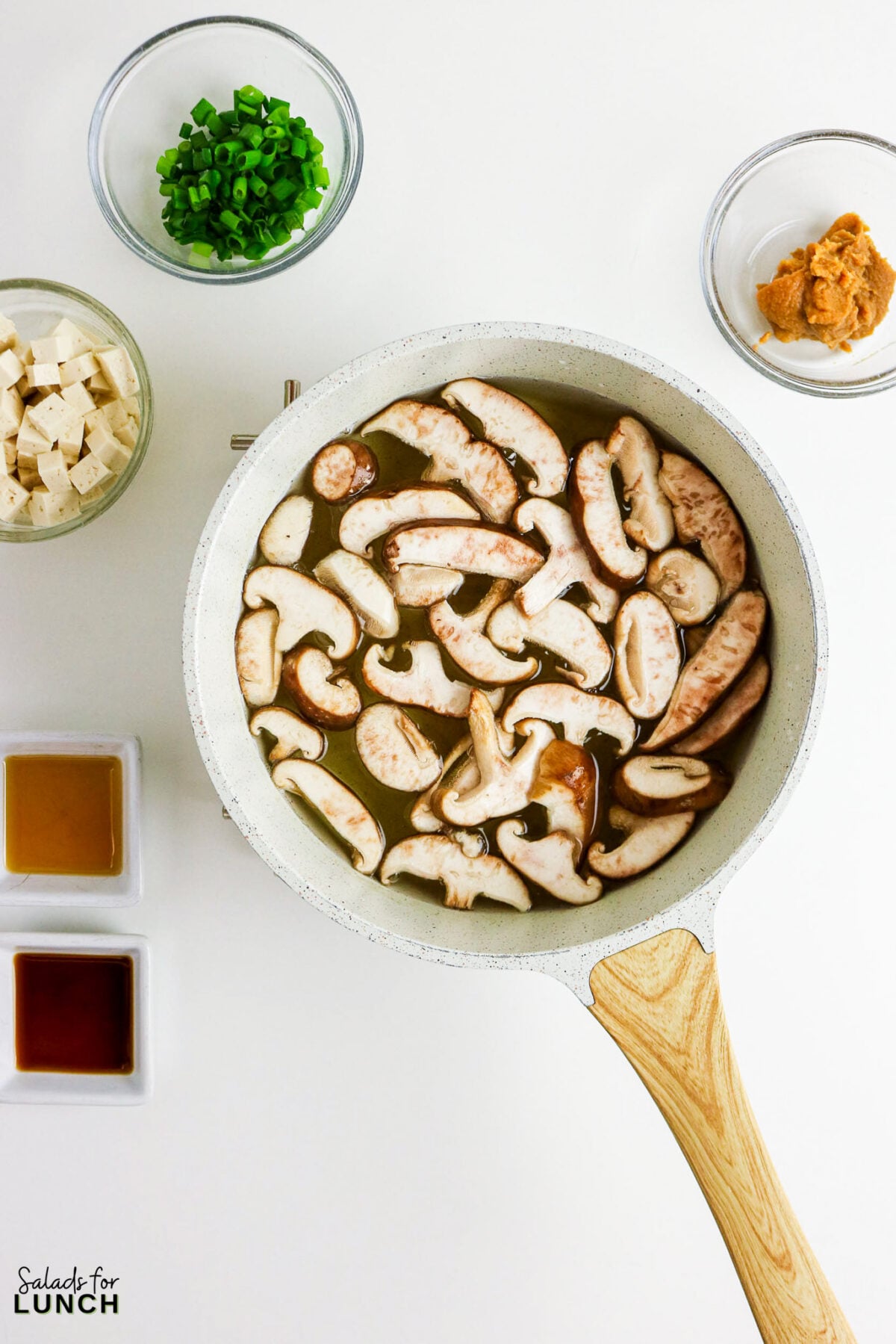Easy Miso Soup with Mushrooms - Miso Soup Recipe With Mushrooms 30 Sliced mushrooms simmering in dashi broth in a saucepan, surrounded by bowls of cubed tofu, green onions, miso paste, soy sauce, and sesame oil.