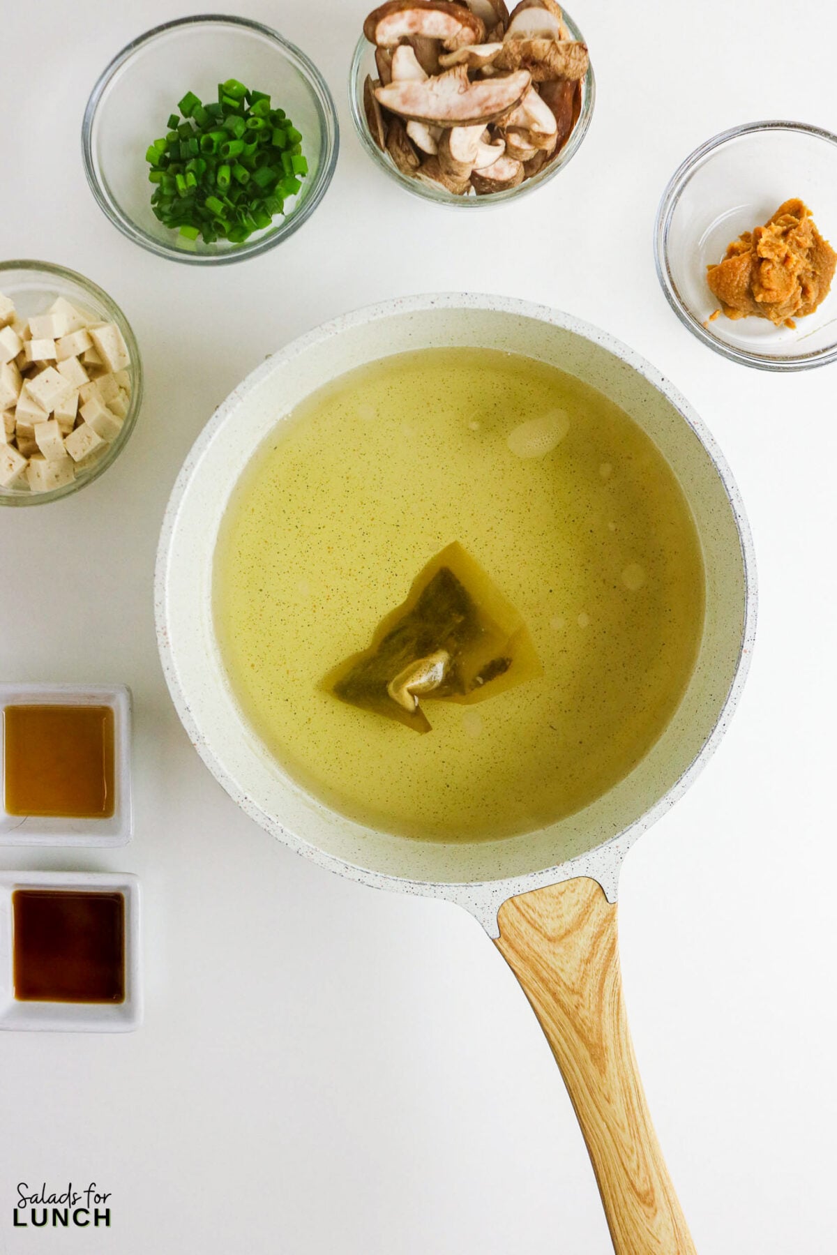 Easy Miso Soup with Mushrooms - Miso Soup Recipe With Mushrooms 31 Dashi packet simmering in a saucepan surrounded by prepped ingredients: tofu cubes, sliced mushrooms, green onions, miso paste, soy sauce, and sesame oil.