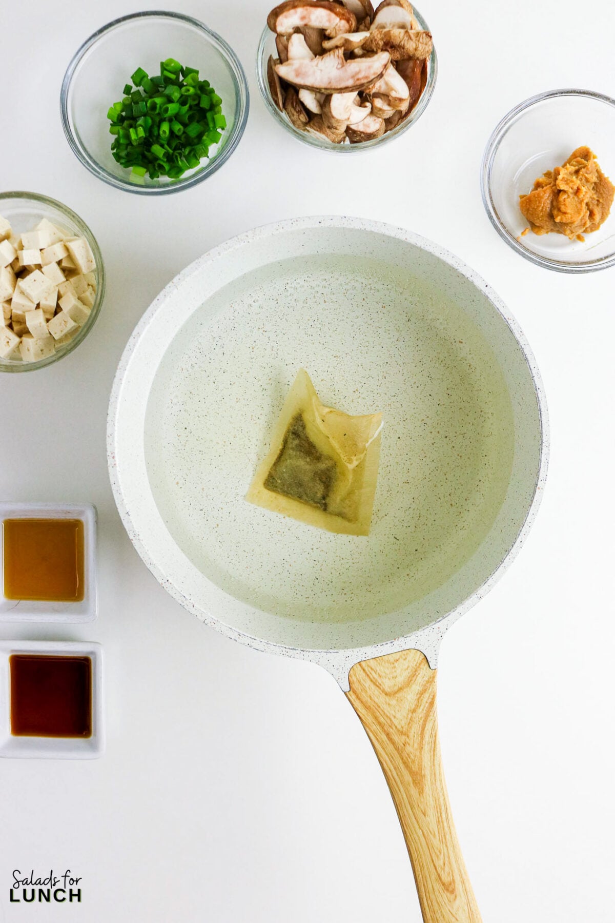 Easy Miso Soup with Mushrooms - Miso Soup Recipe With Mushrooms 32 Dashi packet simmering in water in a saucepan, surrounded by bowls of tofu, mushrooms, green onions, miso paste, soy sauce, and sesame oil.