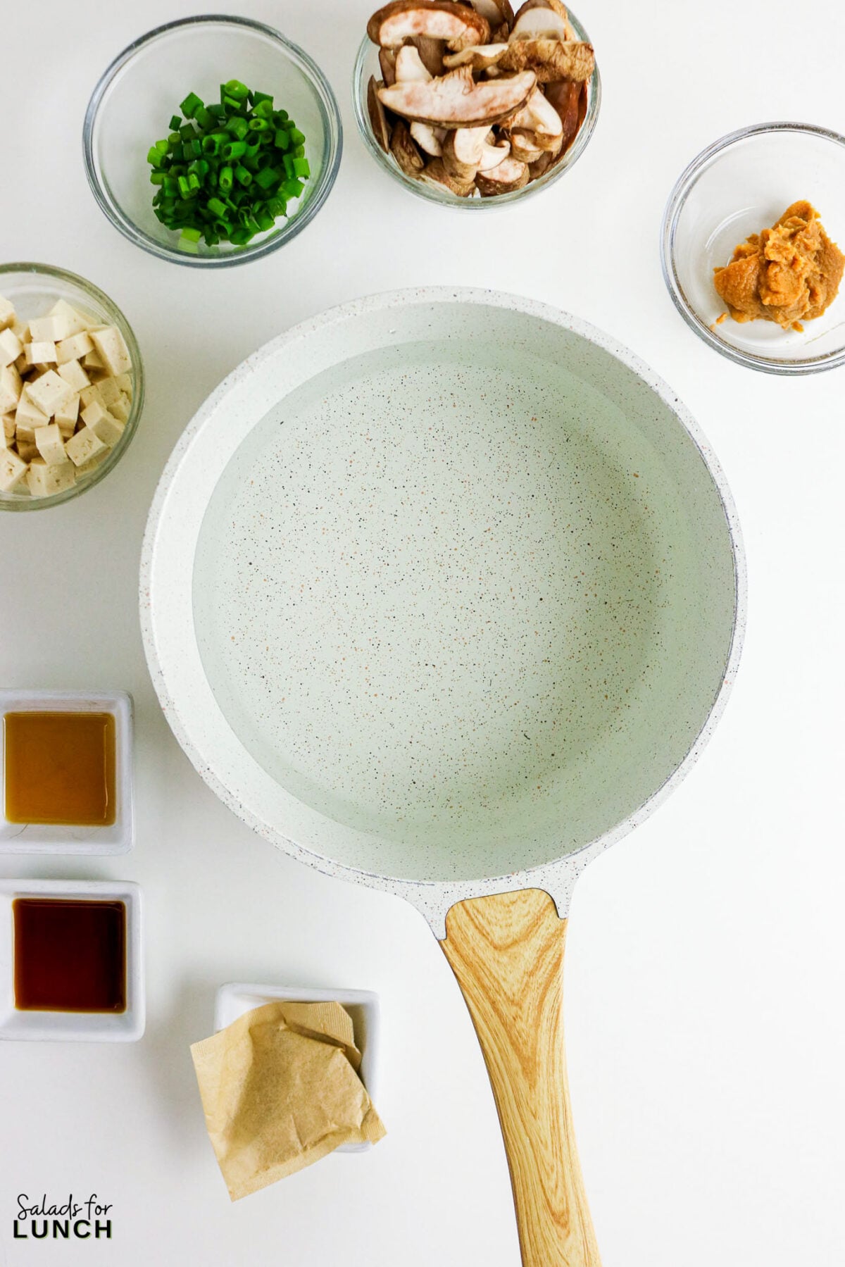 Easy Miso Soup with Mushrooms - Miso Soup Recipe With Mushrooms 33 Miso soup ingredients laid out around an empty saucepan: tofu cubes, sliced mushrooms, green onions, miso paste, soy sauce, sesame oil, and a dashi packet.