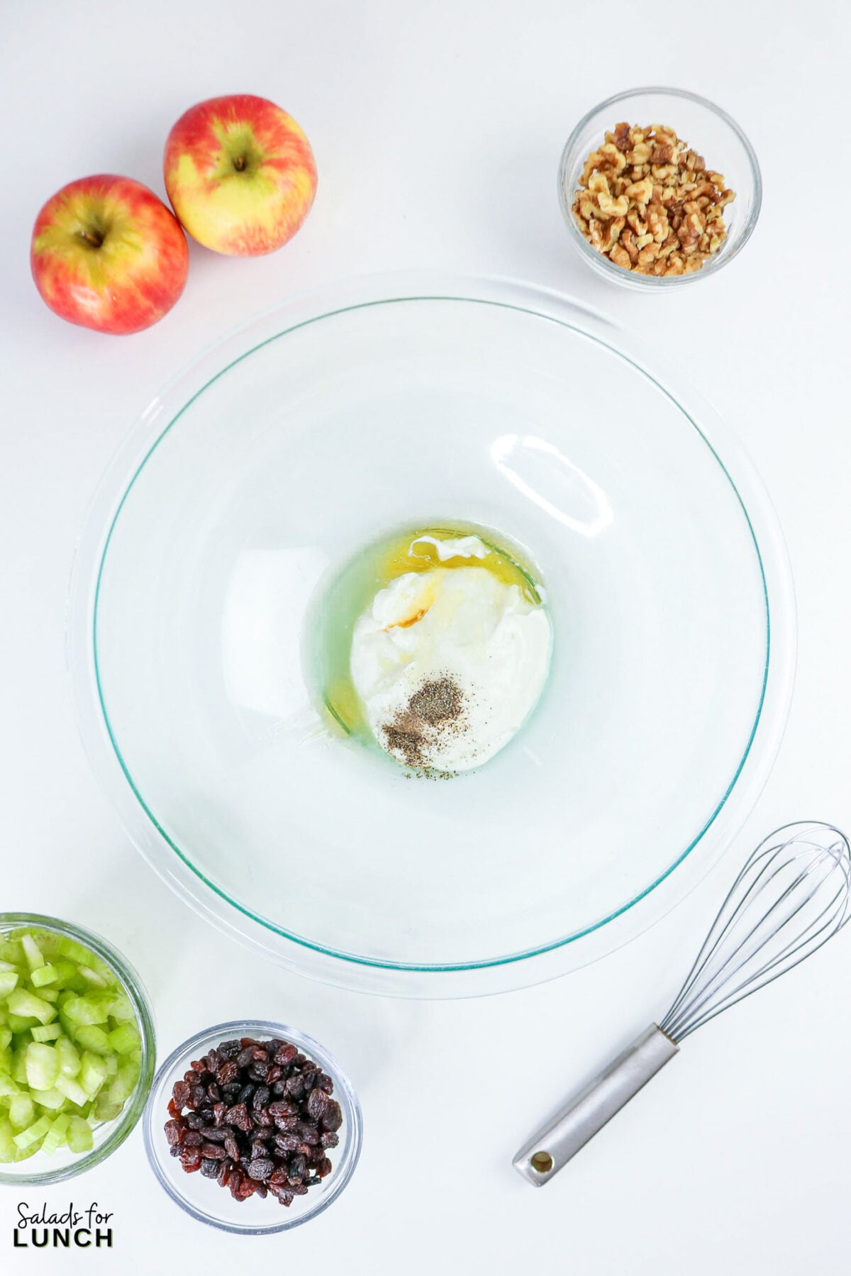 Apple, Celery & Walnut Crunch Salad - Process Apple Celery Walnut Salad 4 Step 1 of Healthy Waldorf salad ingredients laid out: apples, walnuts, raisins, celery, and dressing in a glass bowl with a whisk.