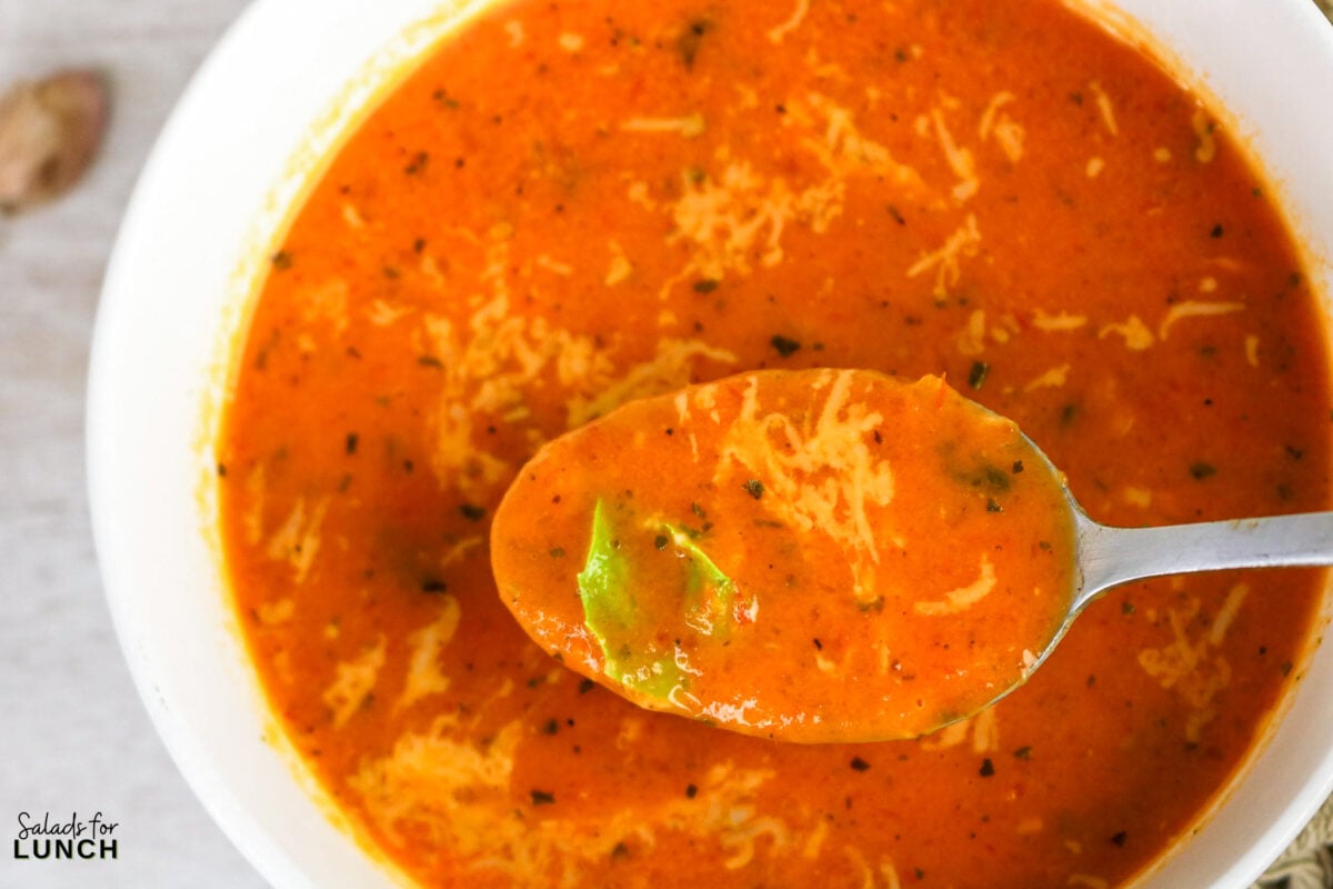 A spoonful of roasted tomato basil soup held over a white bowl, topped with shredded Parmesan and a fresh basil leaf.