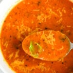 A spoonful of creamy roasted tomato soup topped with shredded cheese and fresh basil held over a full bowl of the same soup.