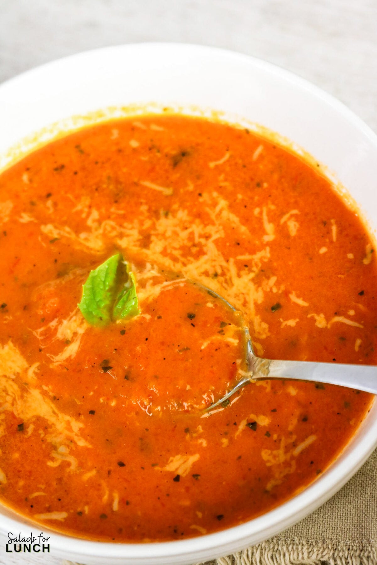 A white bowl of creamy tomato soup topped with shredded cheese and a fresh basil leaf, with a spoon resting in it.