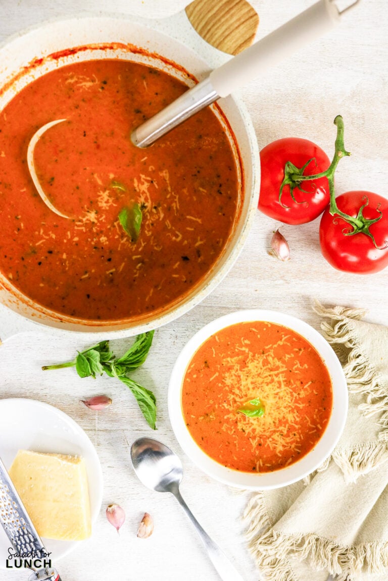 Overhead view of tomato soup topped with shredded Parmesan and fresh basil, served in a bowl and a large pot, with tomatoes, garlic, and cheese nearby.