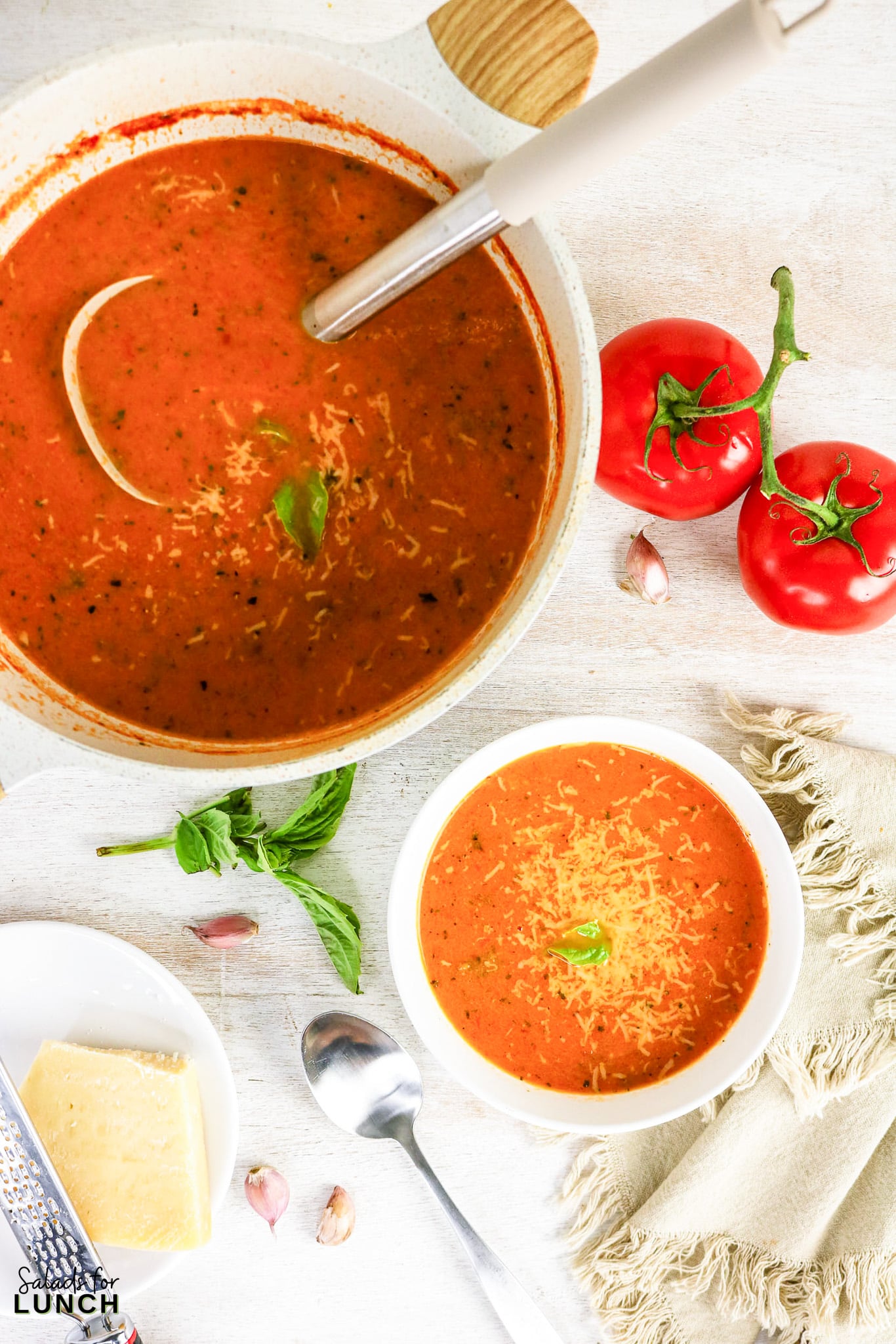 Overhead view of tomato soup topped with shredded Parmesan and fresh basil, served in a bowl and a large pot, with tomatoes, garlic, and cheese nearby.