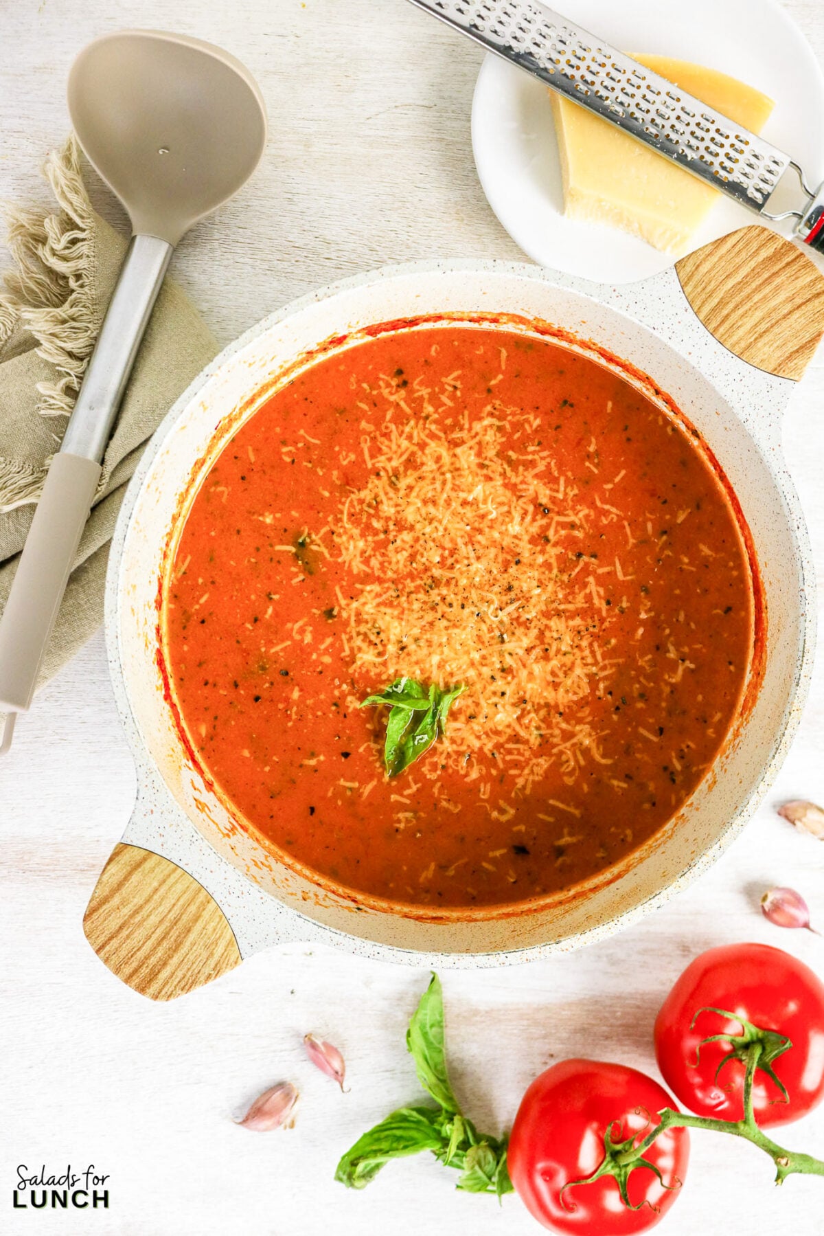 Roasted tomato basil soup in a white pot, topped with grated Parmesan and fresh basil, surrounded by tomatoes, garlic, and a block of Parmesan.