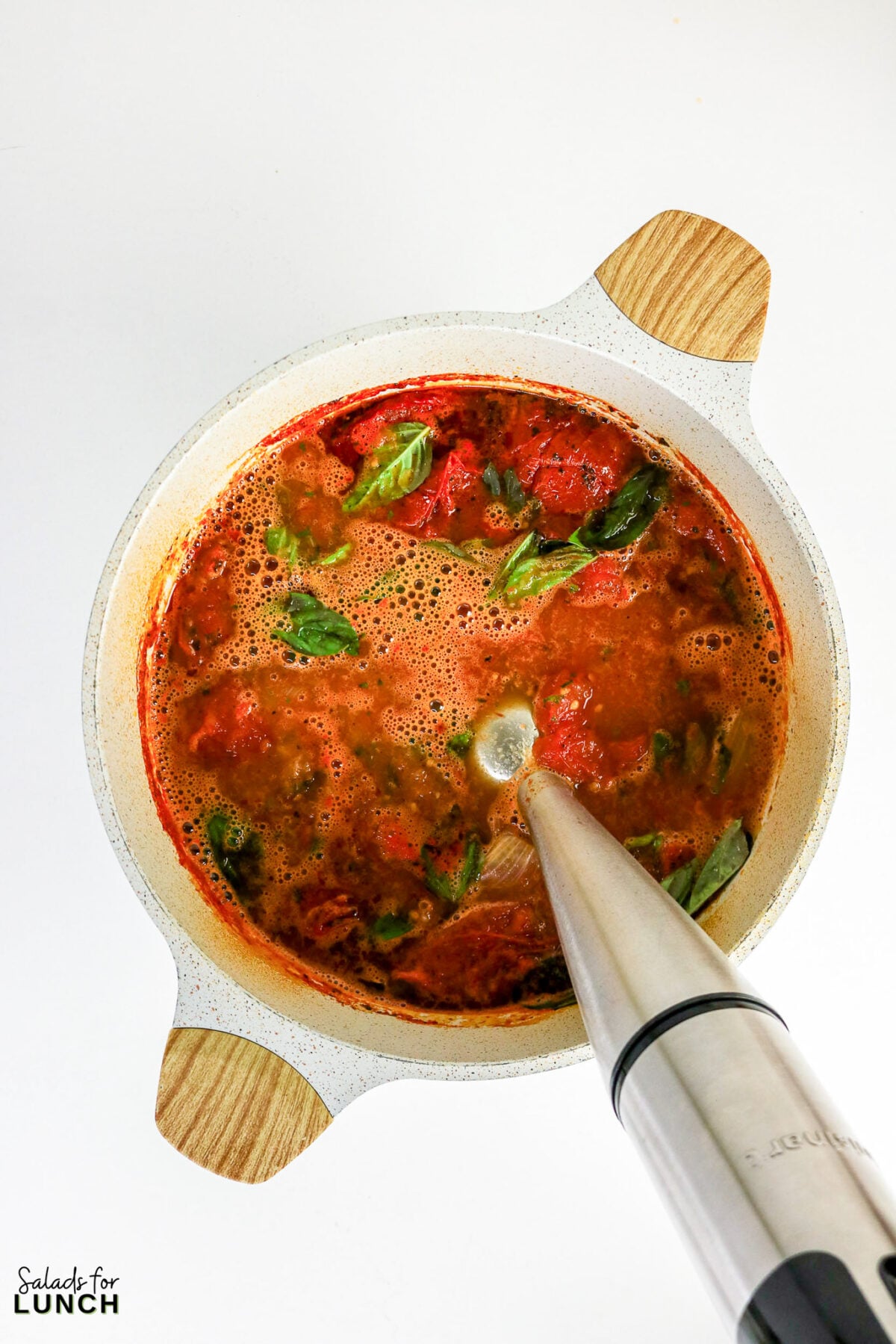 Immersion blender blending roasted tomatoes and fresh basil leaves directly in a large pot.