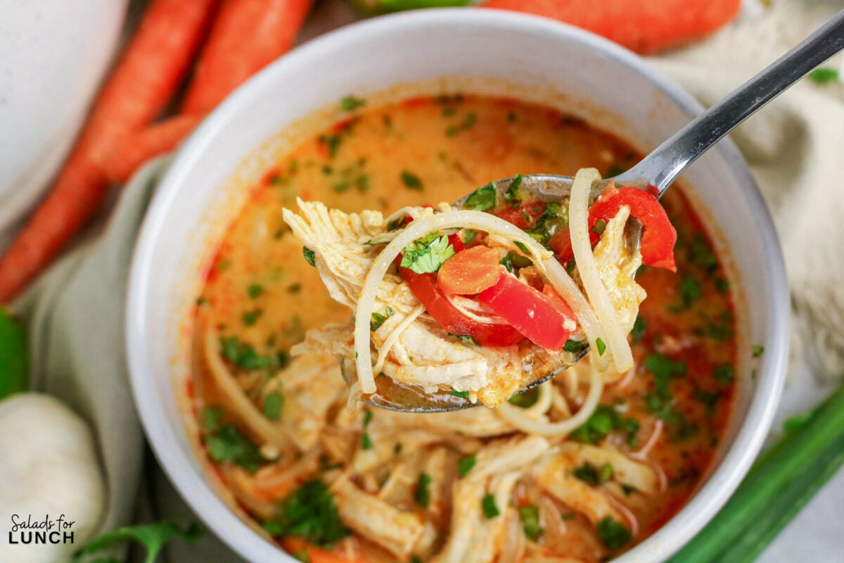 Spoonful of Thai coconut curry soup with shredded chicken, rice noodles, red bell pepper, and cilantro in a creamy broth.
