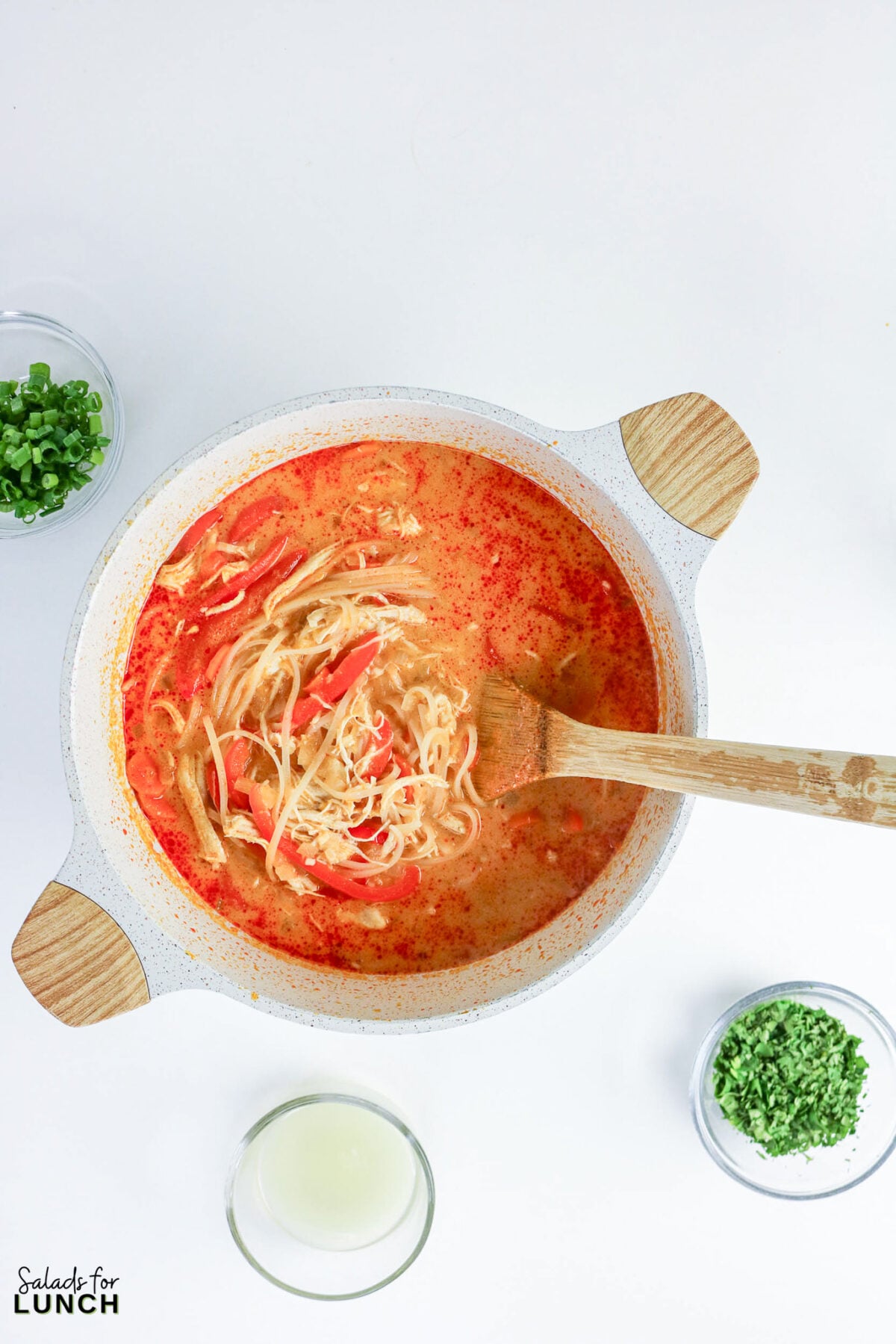 Pot of Thai coconut curry soup with rice noodles, shredded chicken, and bell pepper, with lime juice and herb garnishes on the side.
