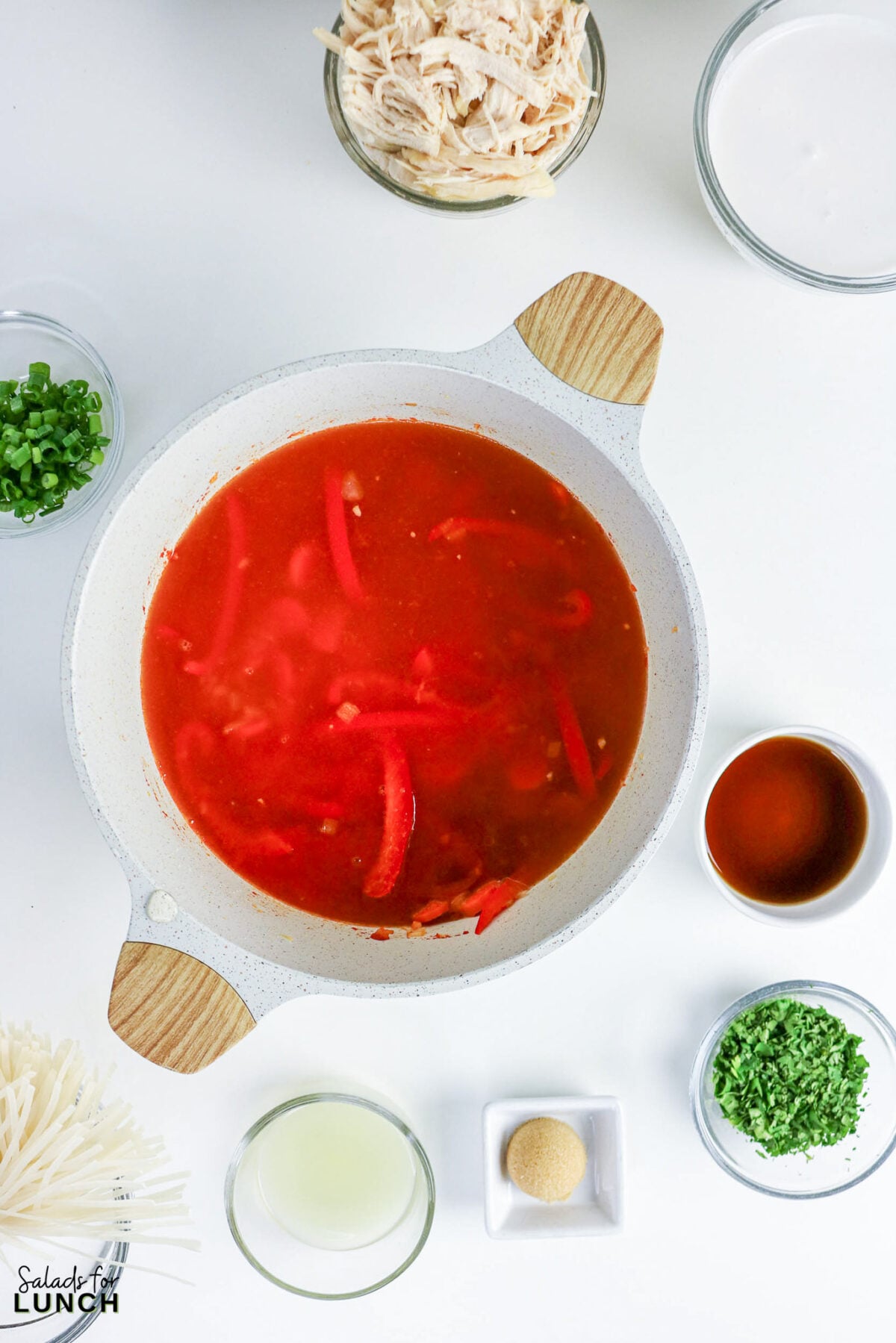 Simmering red curry broth with bell peppers in a pot, surrounded by bowls of shredded chicken, rice noodles, fish sauce, lime juice, and garnishes.