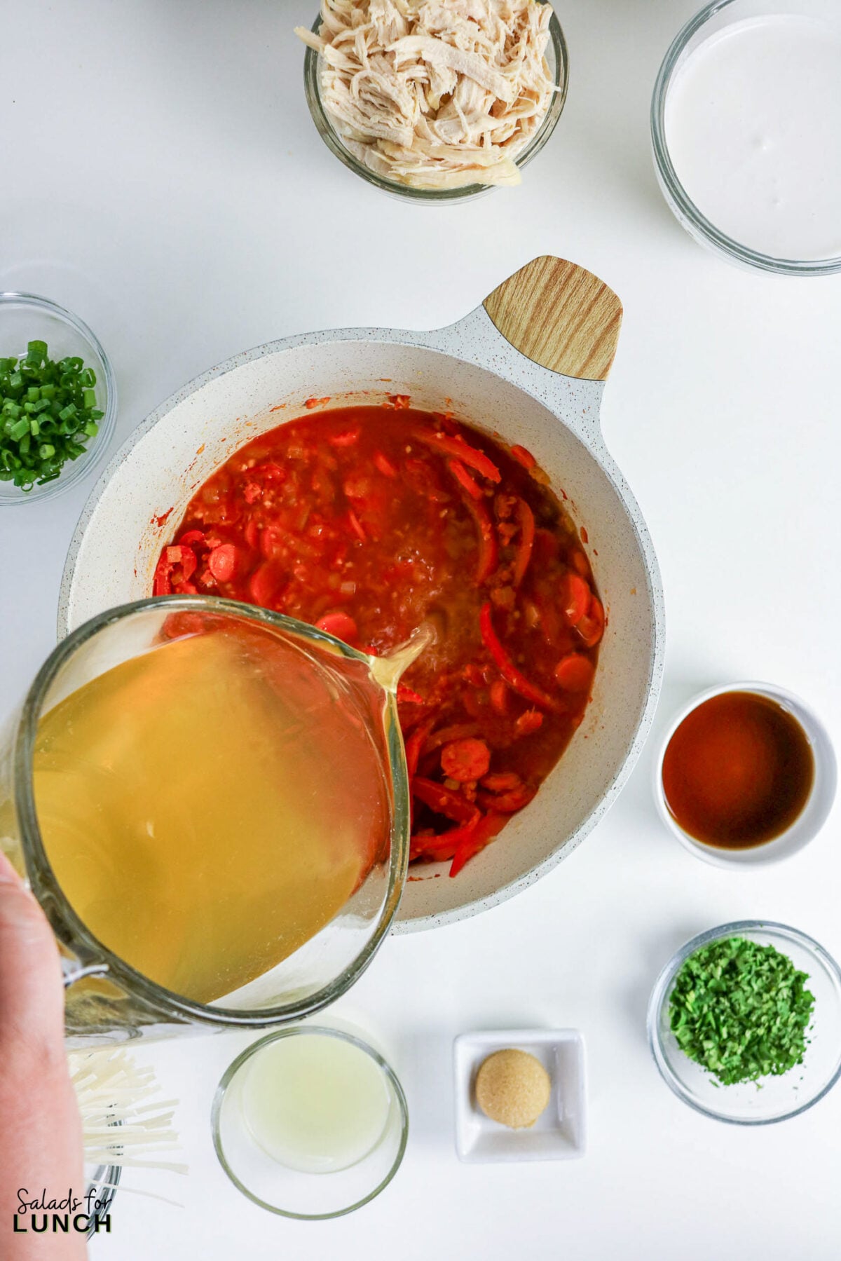 Pouring chicken broth into the pot with sautéed vegetables and curry paste, with bowls of coconut milk, chicken, and garnishes nearby.