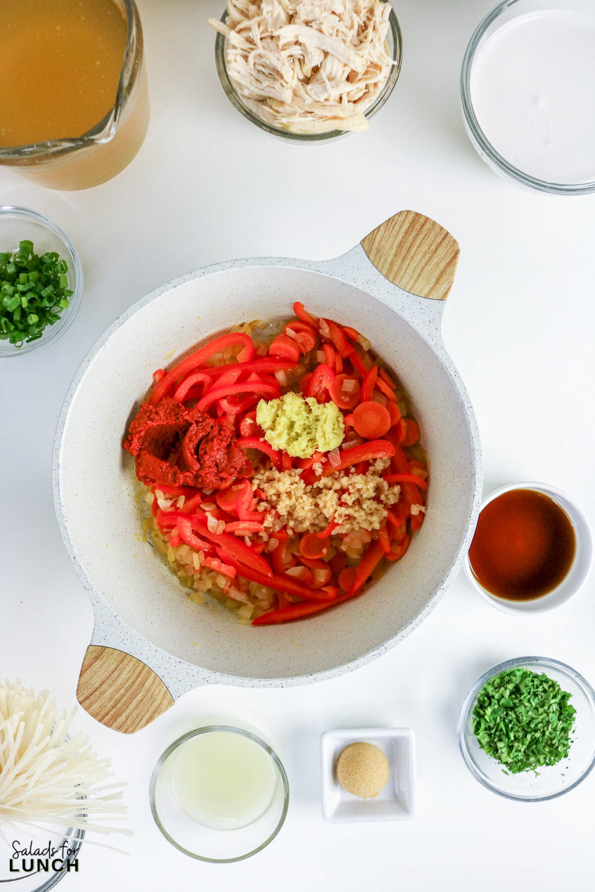 Pot with sautéed vegetables, garlic, ginger, and red curry paste, surrounded by bowls of broth, coconut milk, shredded chicken, and other soup ingredients.