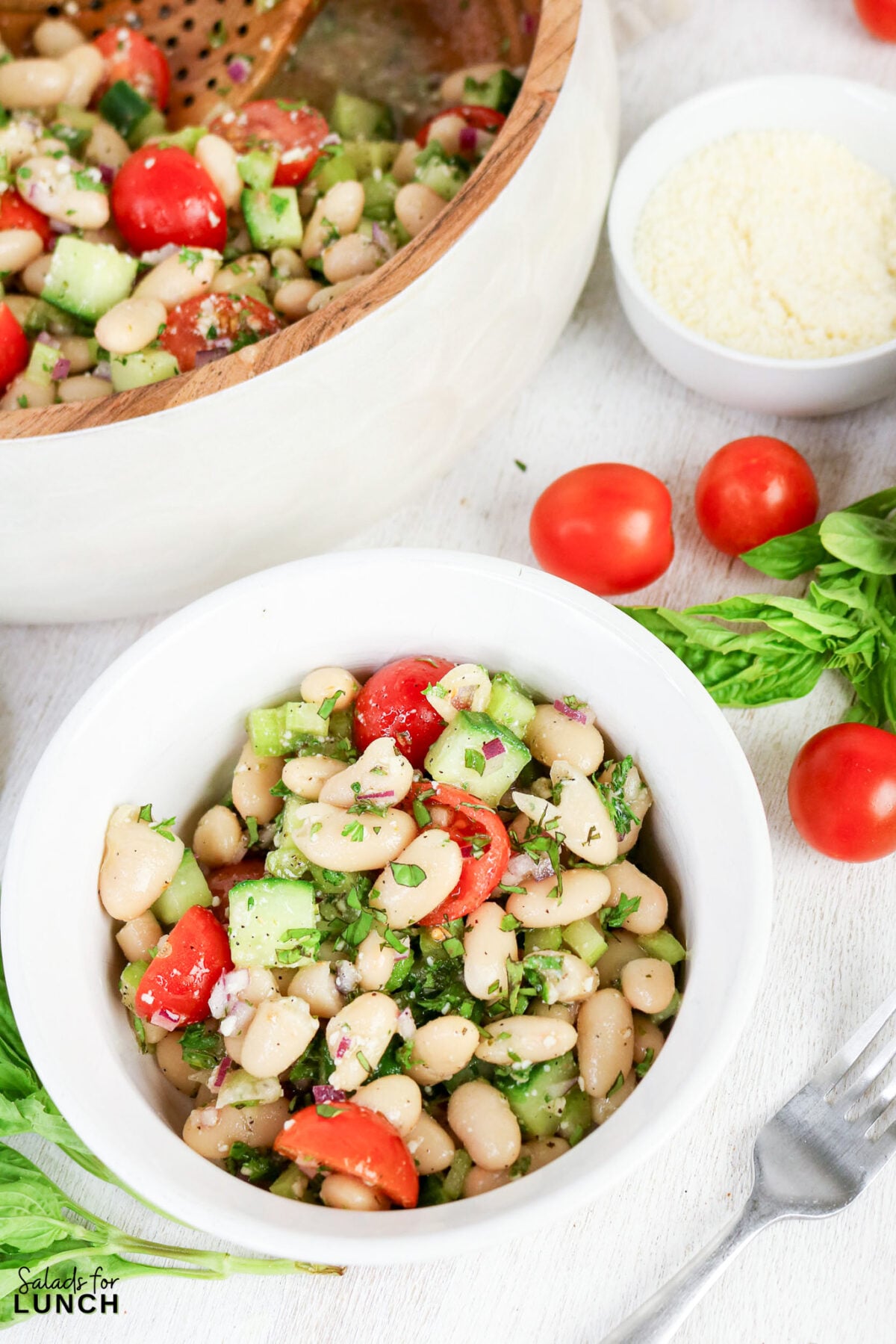 Easy Tuscan Bean Salad - Tuscan Bean Salad Hero 07 White bean Tuscan salad with cherry tomatoes, cucumber, red onion, and fresh herbs served in a white bowl, with a larger serving bowl behind it.