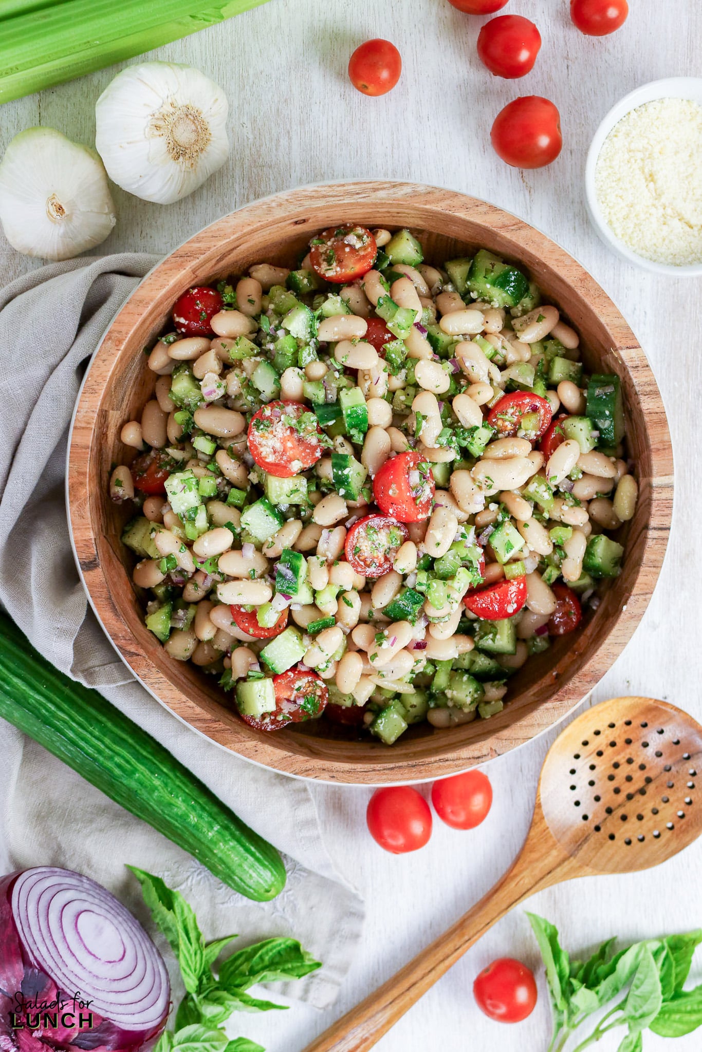 White bean salad with cherry tomatoes, cucumber, and herbs in a wooden bowl, surrounded by fresh ingredients.